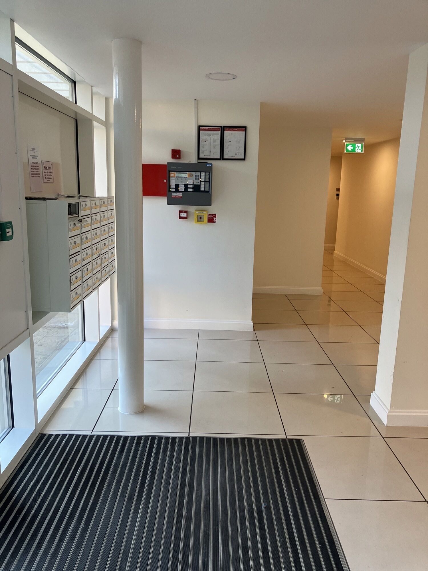 A clean, modern lobby with a tiled floor, a black and white striped mat, a fire alarm panel, and a red emergency exit sign.