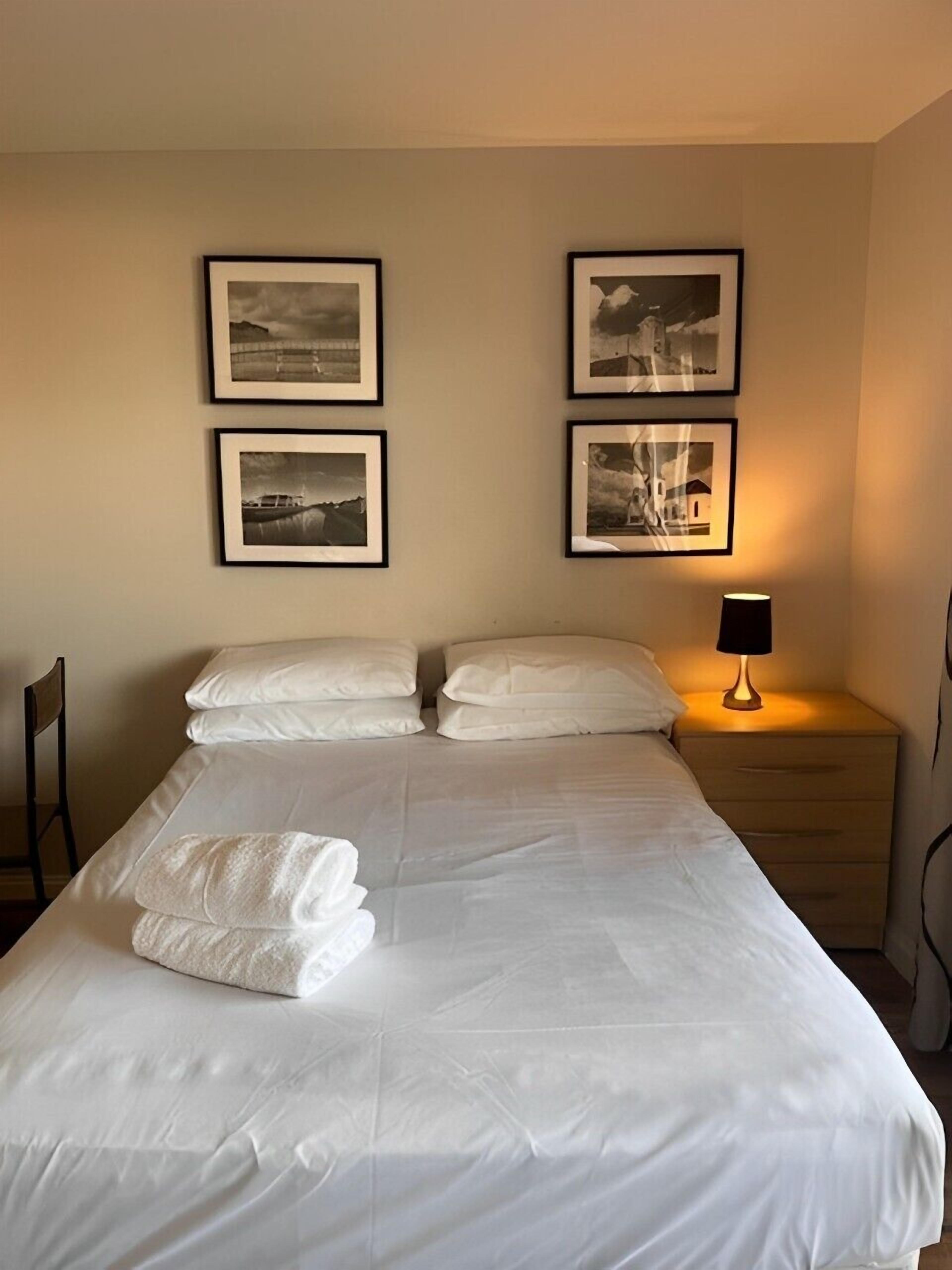 A neatly made bed with white linens and towels, a bedside lamp, and three framed pictures on the wall.