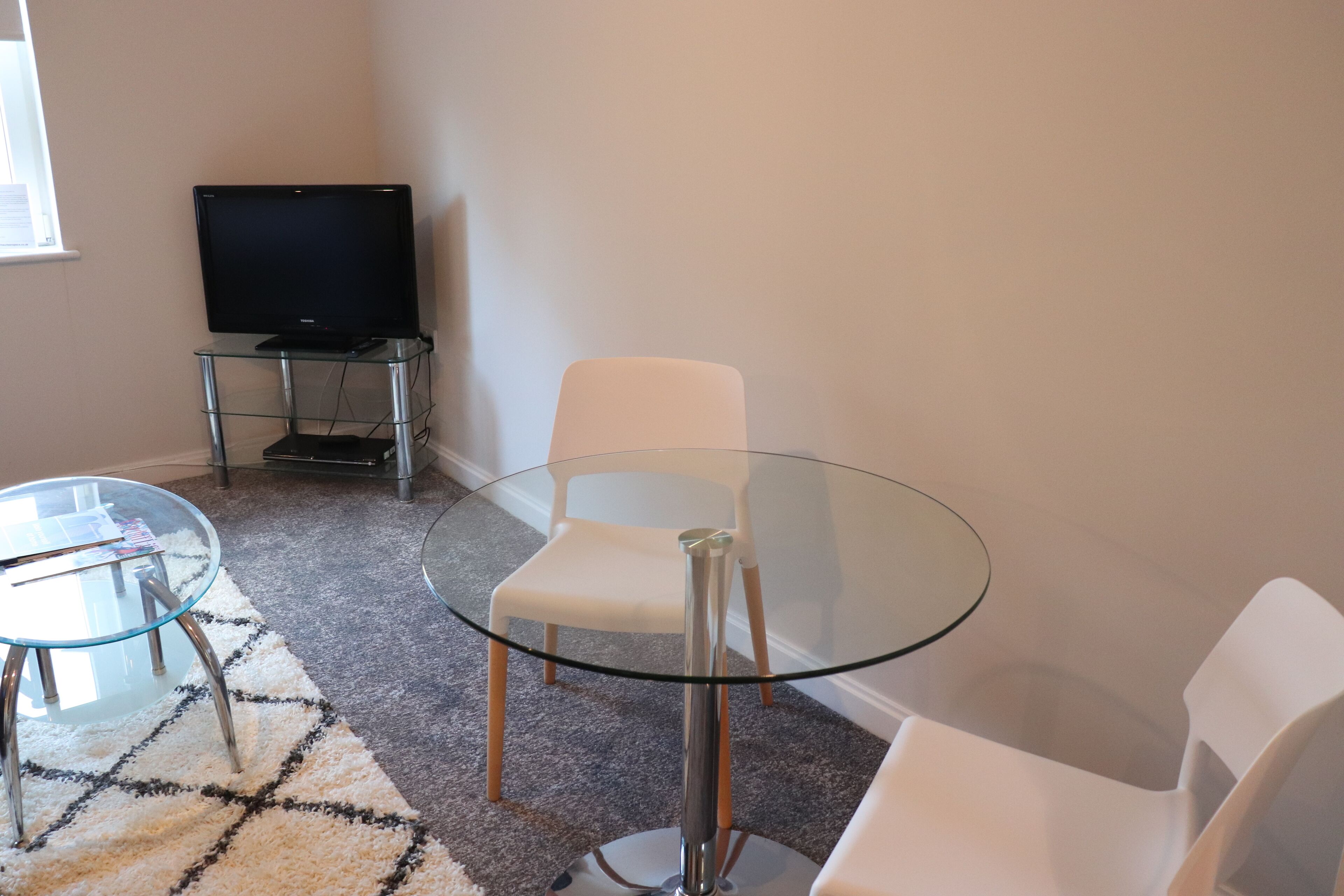Un salón moderno con una mesa de vidrio, sillas blancas, un televisor de pantalla plana sobre un mueble bajo y suelo alfombrado.