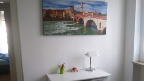 A bedroom with a bed, a desk with a chair, a lamp, a painting of a bridge, and a window. - Quiet room in new apartment (Verona)