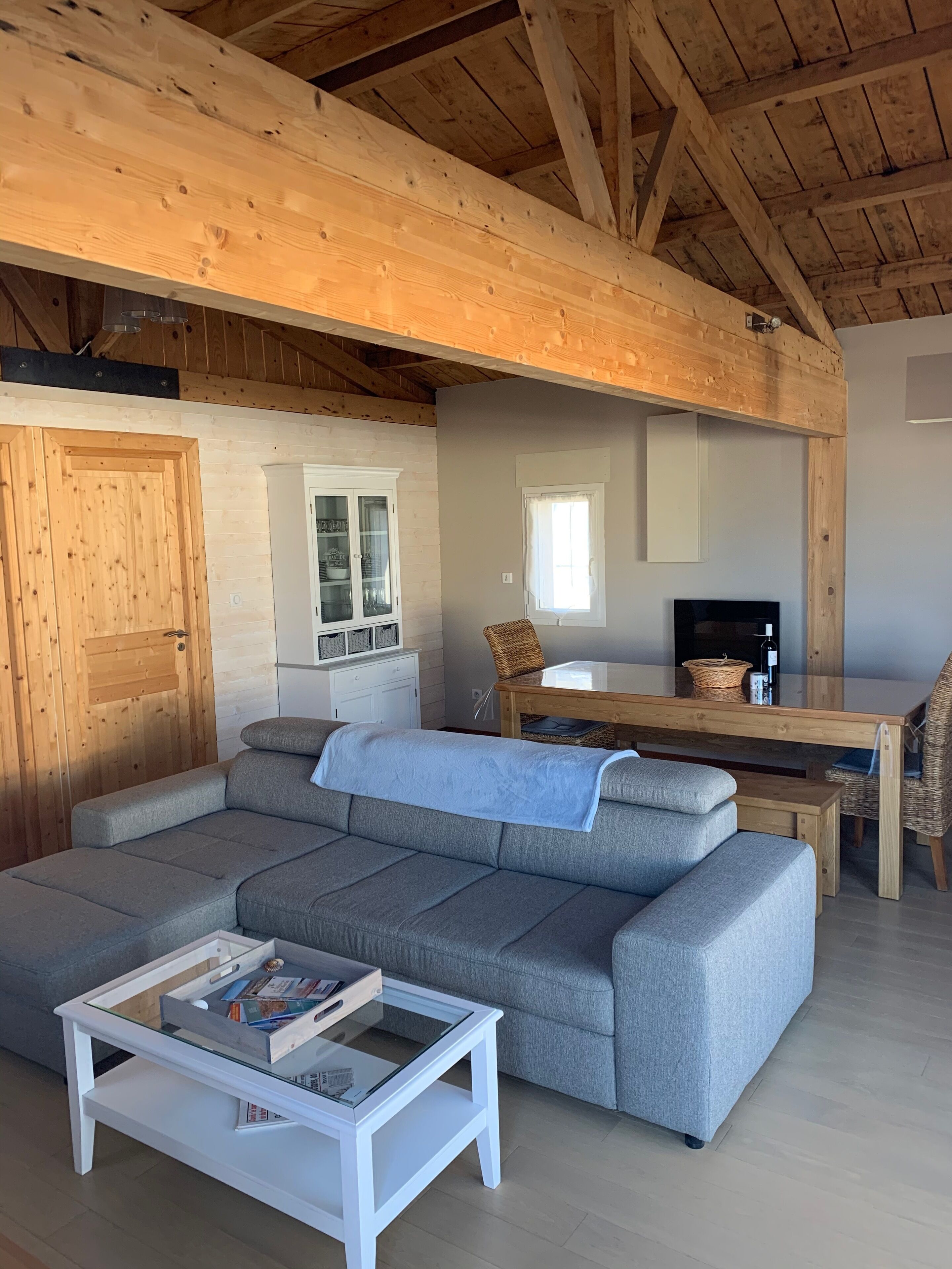 Een gezellige woonkamer met een grijze modulaire bank, een witte salontafel met boeken, een houten eettafel met een rieten stoel, en een televisie.