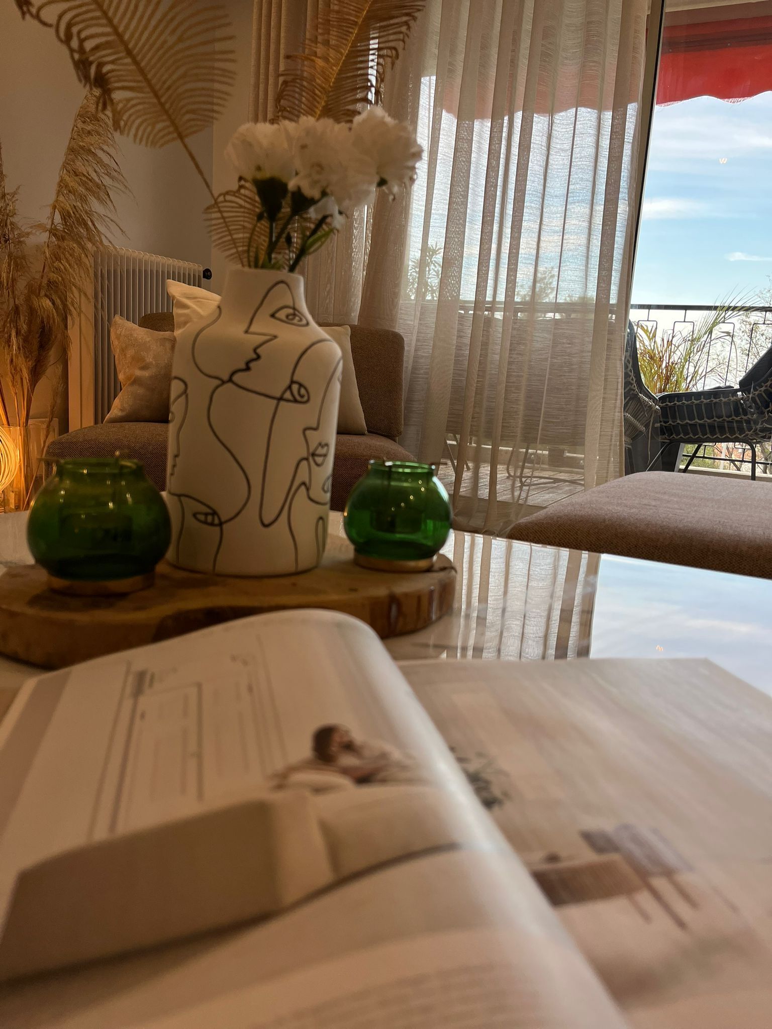 A cozy living room with a sofa, a coffee table with a book, decorative vases, and a view of the outdoors through sheer curtains.