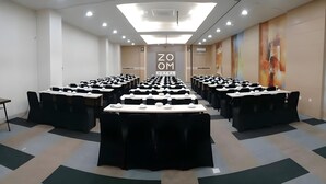 Uma sala de conferências com várias mesas e cadeiras, teto com iluminação embutida e uma parede com um letreiro grande escrito 'ZOOM HOTEL'.