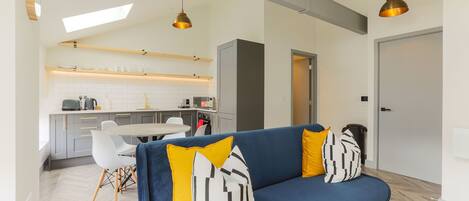 A modern living room with a blue sofa, yellow and black patterned pillows, a dining table with chairs, and a kitchen in the background.