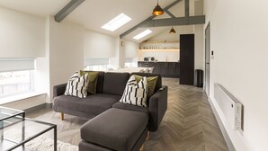 A modern living room with a grey sectional sofa, ottoman, and glass coffee table. A kitchen is visible in the background with white cabinets and a black countertop. - Goodstay Apartments by Urban Space (Barry)