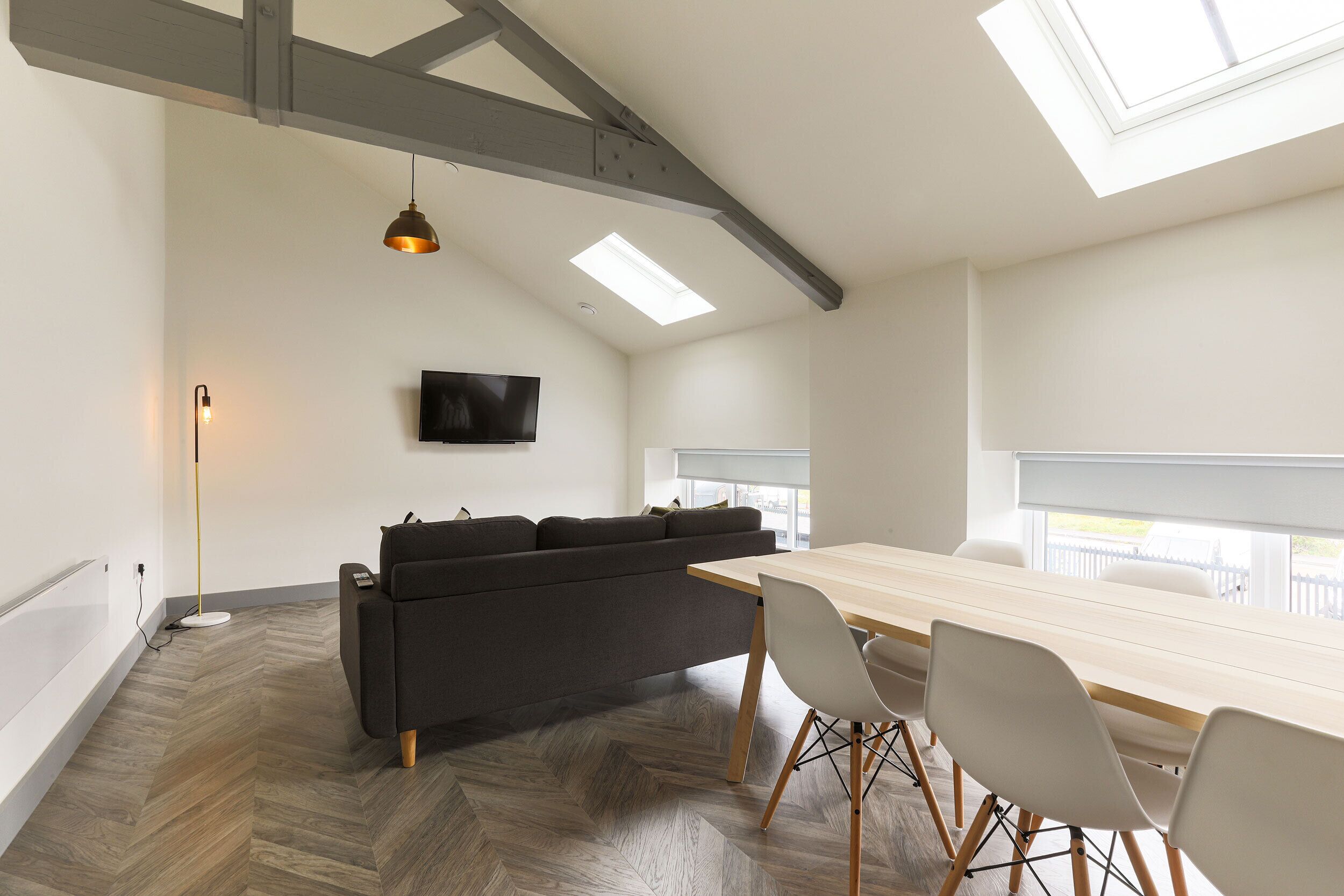 A modern room with a herringbone floor, a dark grey sofa, a wooden dining table with white chairs, a flat-screen TV mounted on the wall, and skylights.