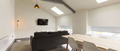 A modern room with a herringbone floor, a dark grey sofa, a wooden dining table with white chairs, a flat-screen TV mounted on the wall, and skylights.