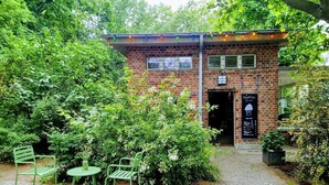 A brick building with a small entrance, surrounded by greenery and a paved area with two green chairs and a table. - Quiet studio in Prenzlauer Berg near Mauerpark (Berlin)