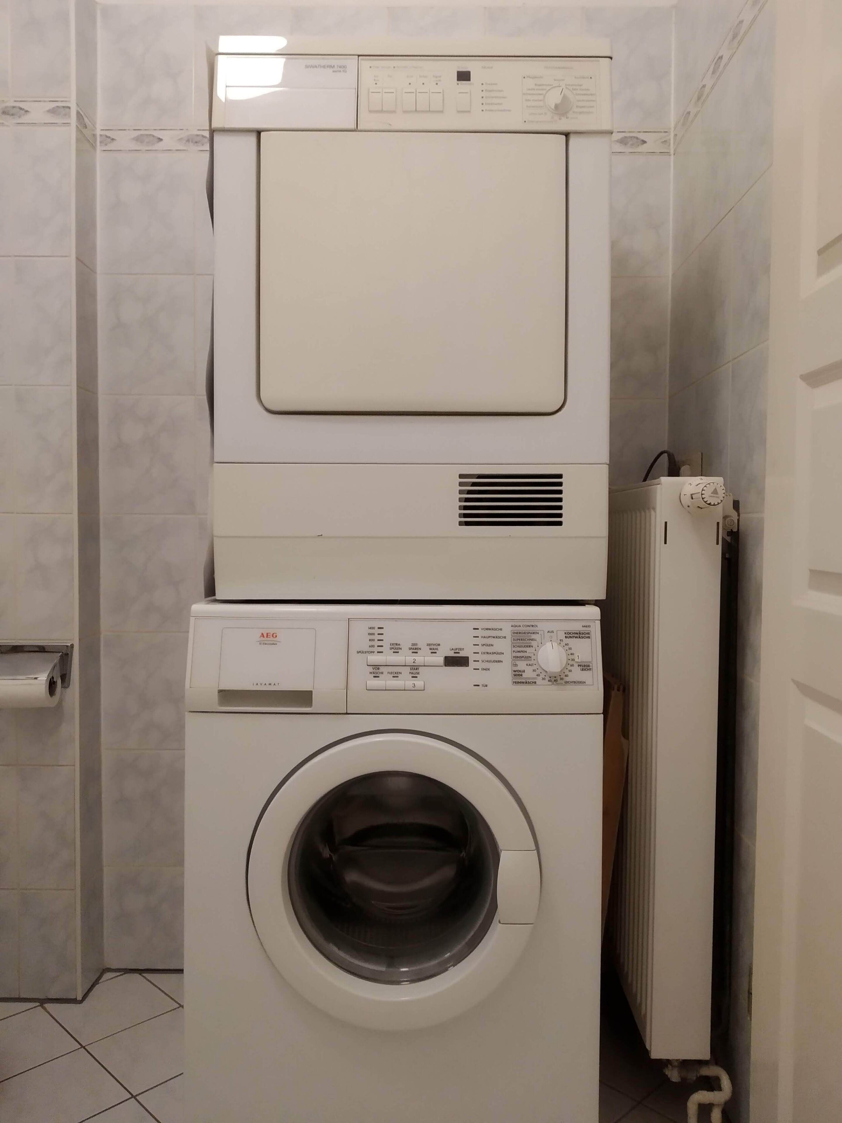 Eine gestapelte Waschmaschine und ein Trockner in einem Badezimmer mit Fliesenwänden und einem sichtbaren Toilettenpapierhalter.