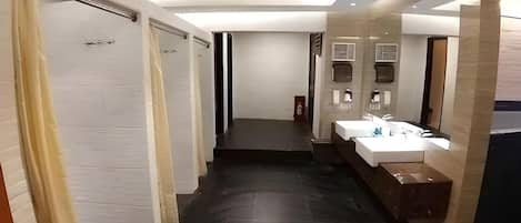 A modern hotel corridor with a central staircase, a bathroom with a large mirror and sink, and a fire extinguisher on the wall.