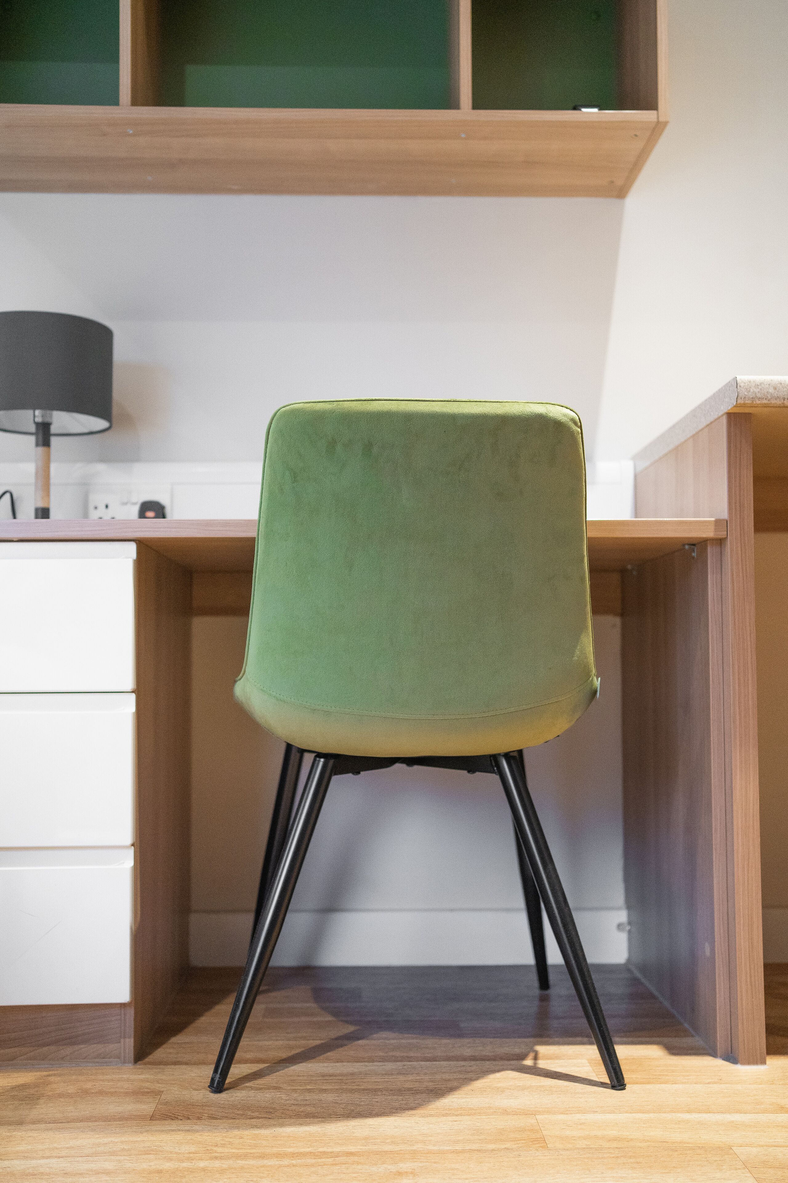 Una sedia verde con gambe nere in una stanza con pavimento in legno e una scrivania bianca.