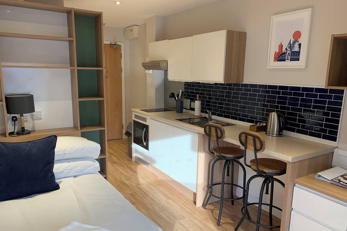 A compact hotel room with a kitchenette, a bed with white linens, a desk with a lamp, and a framed picture on the wall.