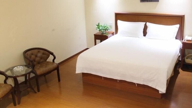 A hotel room with a wooden bed, a small round table, two chairs, a nightstand with a plant, and a painting on the wall. - Greentree Inn Xuzhou West District Huohua Shell Ho (Xuzhou)