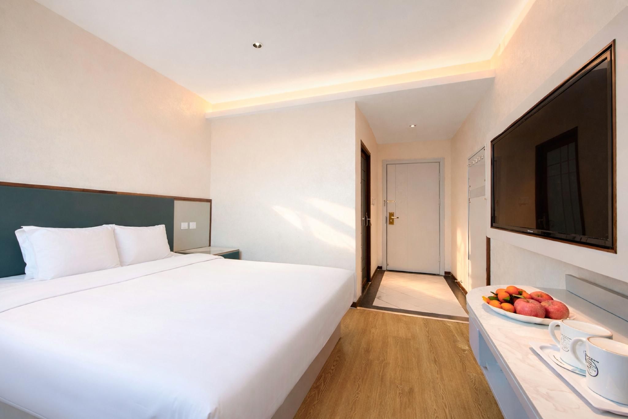 A hotel room with a bed, a bedside table, a mirror, a door, and a small kitchenette area with fruit and a cup.