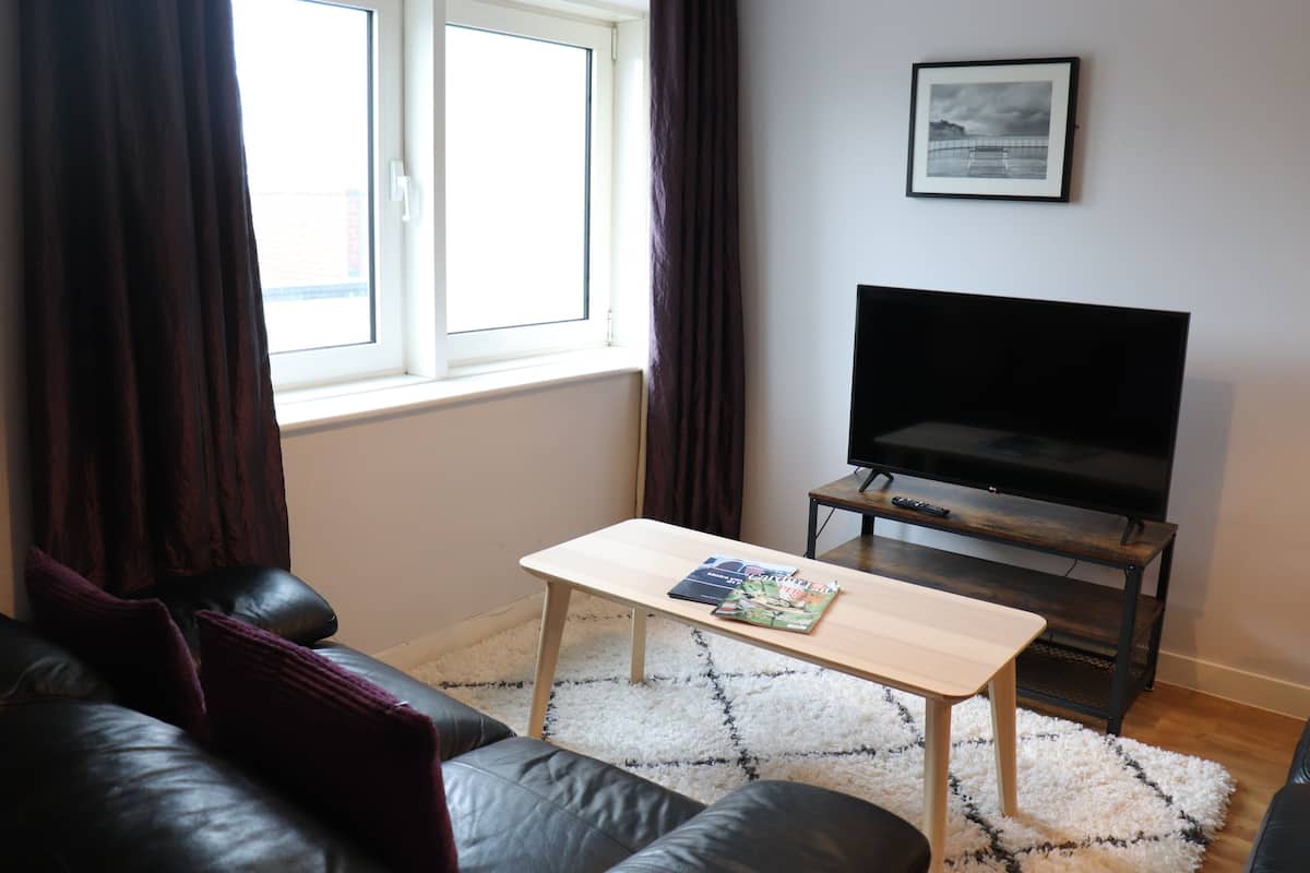 A living room with a leather sofa, a wooden coffee table with magazines, a TV on a stand, and a window with curtains.