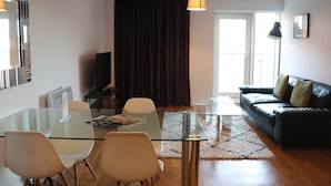 A modern living room with a glass dining table, white chairs, a black leather sofa, and a large window with curtains. - Quayside by Urban Space (Cardiff)