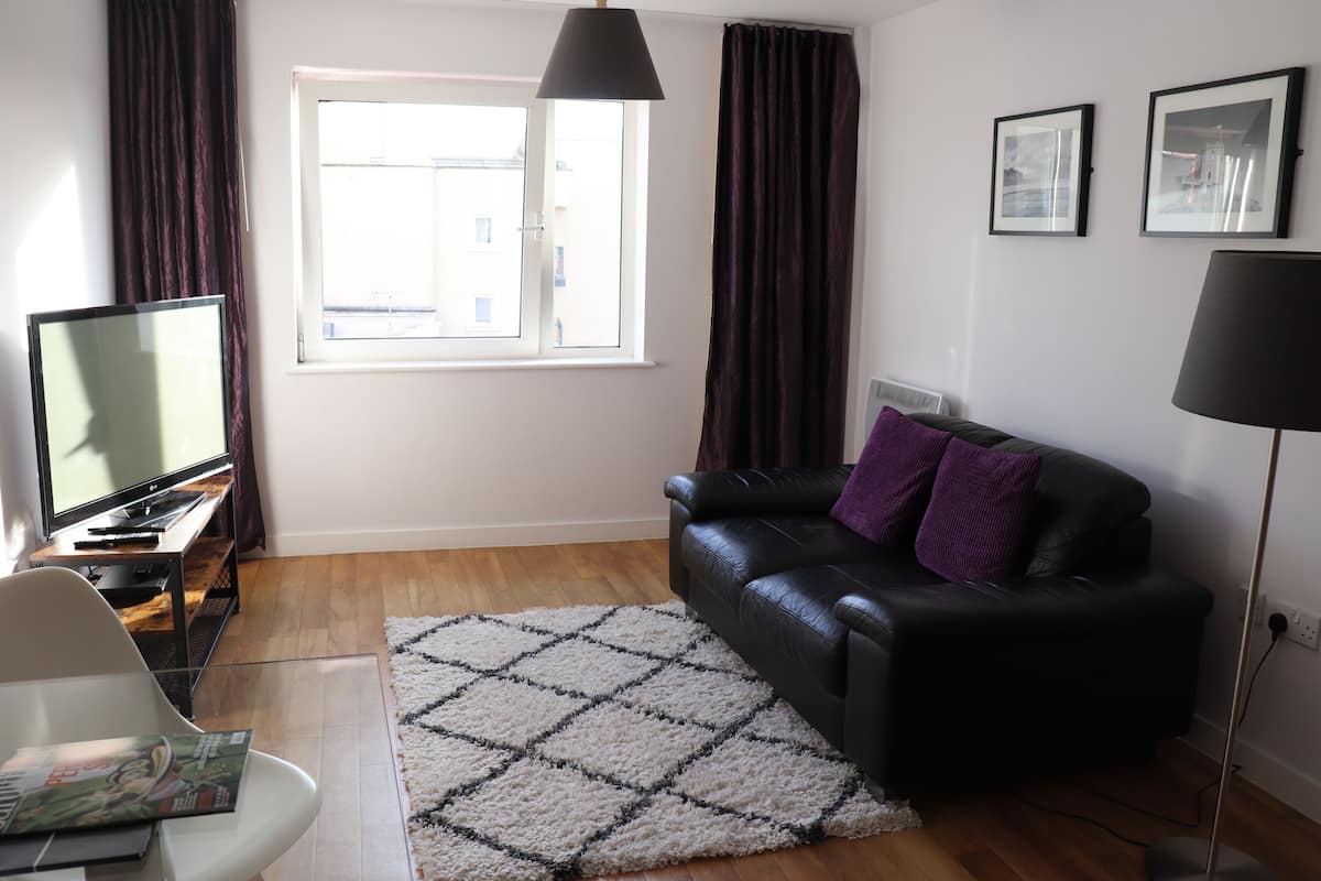 A modern living room with a black leather sofa, a white armchair, a television, and a large window with curtains.