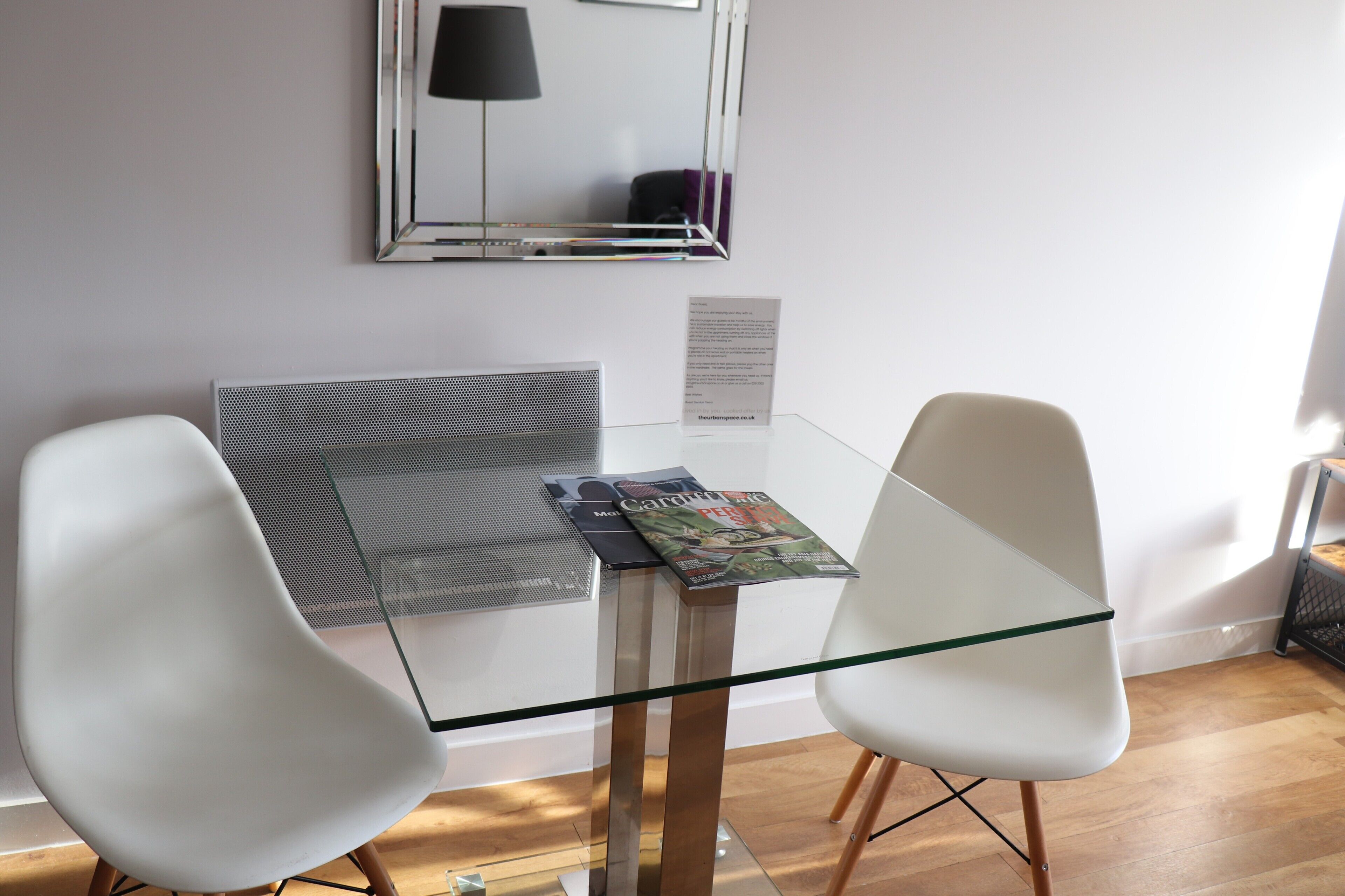 Een moderne kamer met een glazen tafel, twee witte stoelen, een spiegel en een lamp.