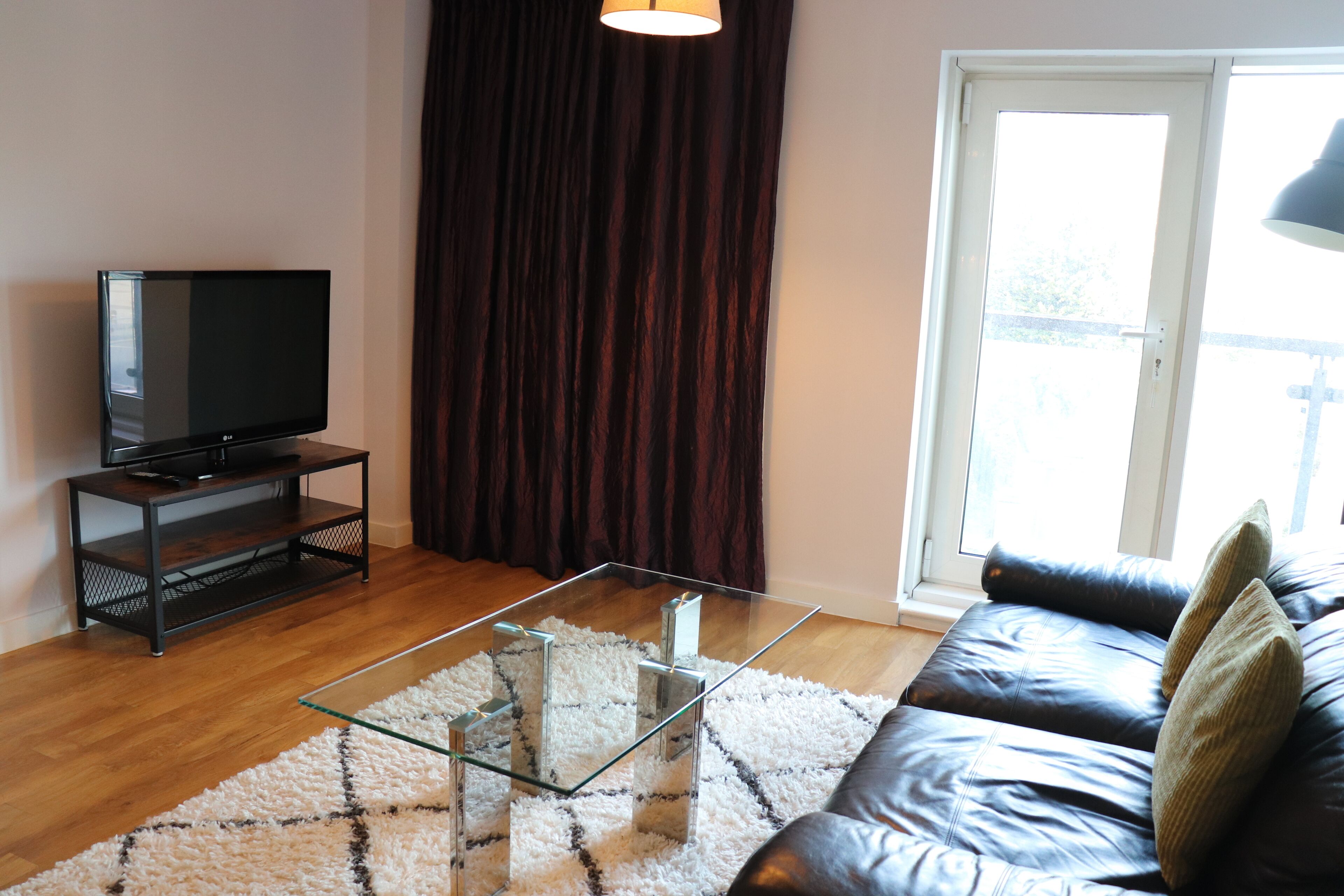 A living room with a flat-screen TV on a stand, a glass coffee table, a dark leather sofa, and a large window with curtains.