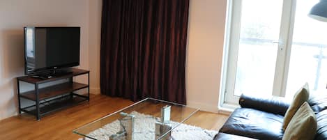Una sala de estar con una televisión de pantalla plana sobre un mueble bajo, una mesa de centro de vidrio, un sofá de cuero oscuro y un ventanal con cortinas.