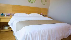 Habitación de hotel con cabecera de madera, cama con ropa blanca, mesita de noche con teléfono, lámpara y una pequeña cesta.
