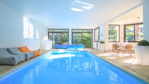 Une piscine intérieure moderne avec des parois en verre transparent, de l’eau bleue et un espace détente avec des coussins.