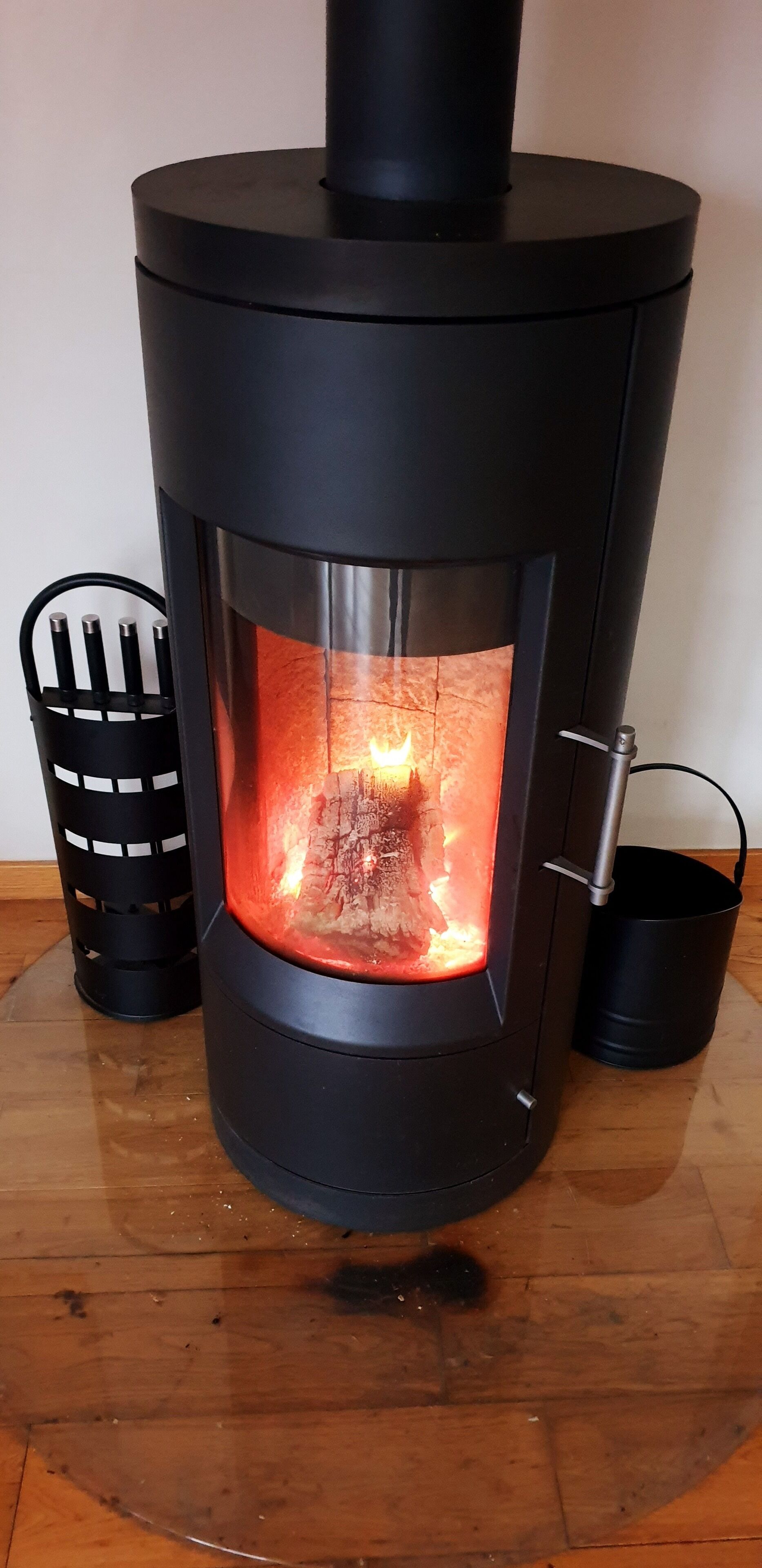 A wood-burning stove with a glass door, a black metal ash collector, and a black metal pot holder.