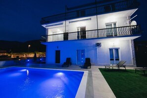 Un edificio de dos pisos con piscina, mobiliario de exterior y un balcón bien iluminado.