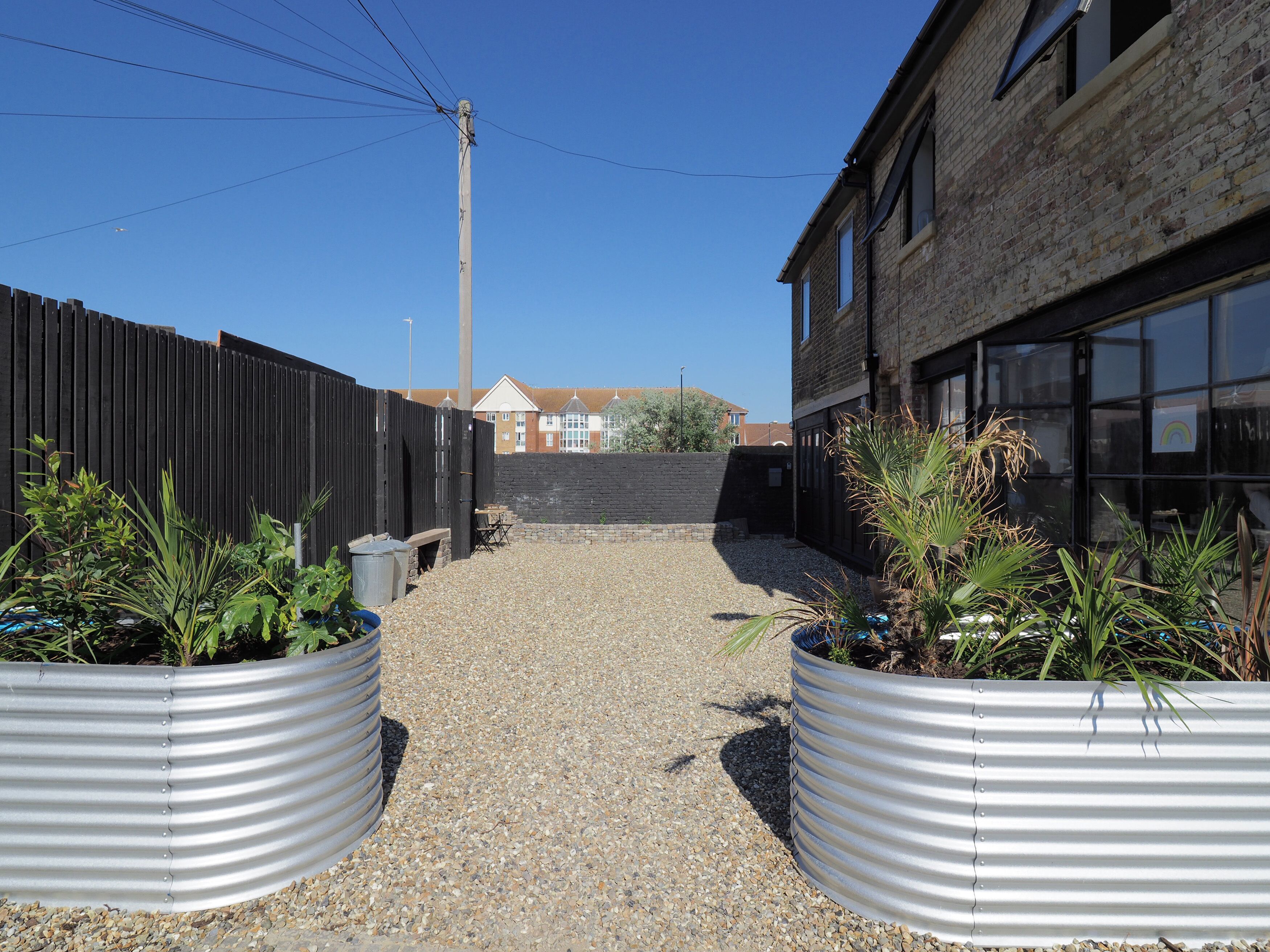 A gravel pathway flanked by metal planters with plants, leading to a building with large windows and a brick facade.