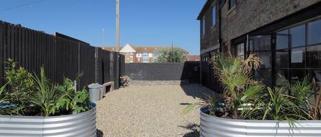 A gravel pathway flanked by metal planters with plants, leading to a building with large windows and a brick facade.
