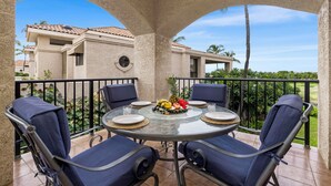 A round glass table with four blue cushioned chairs, a fruit platter, and plates on a balcony with a view of a building and palm trees. - 🏖️Golf Course Views + Large Lanai | Shores #202 (Waikoloa)
