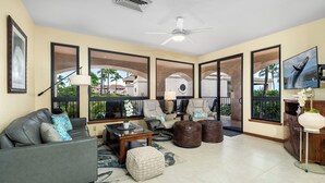 A spacious living room with a gray sofa, a coffee table, and a TV. There are large windows with a view of palm trees and buildings. - 🏖️Golf Course Views + Large Lanai | Shores #202 (Waikoloa)