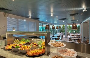 Zona de bufé con diversas frutas y pasteles, diseño interior moderno y amplios ventanales con vistas a la vegetación.