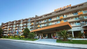 A multi-story hotel with balconies, a central entrance, and a landscaped area with palm trees. - Hotel Excelsior (Golden Sands)