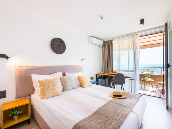 Habitación de hotel con cama, mesita de noche con planta, escritorio con silla y vista al mar desde un balcón.