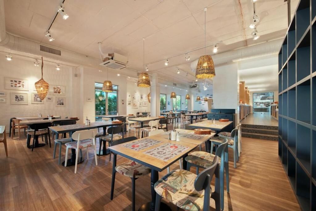 A spacious restaurant with wooden flooring, multiple tables and chairs, hanging wicker lamps, and framed artwork on the walls.