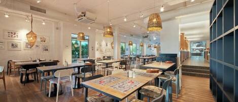 A spacious restaurant with wooden flooring, multiple tables and chairs, hanging wicker lamps, and framed artwork on the walls.