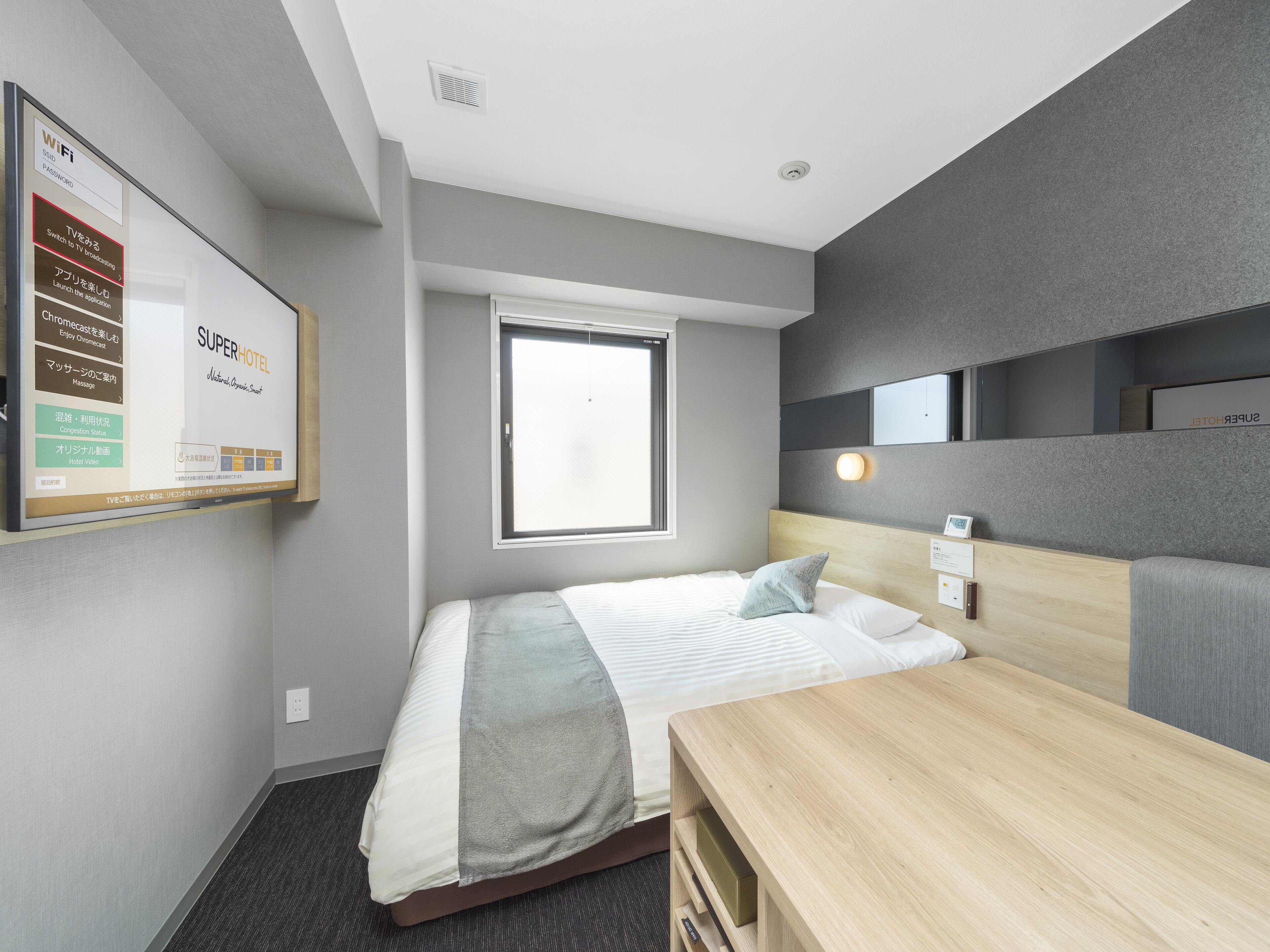A hotel room with a bed, a desk, a window, and a wall-mounted TV displaying hotel amenities.