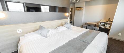 A hotel room with a large bed, a desk, a chair, a small table, and a mirror. There is an air conditioning unit on the wall and a window with a view of buildings.