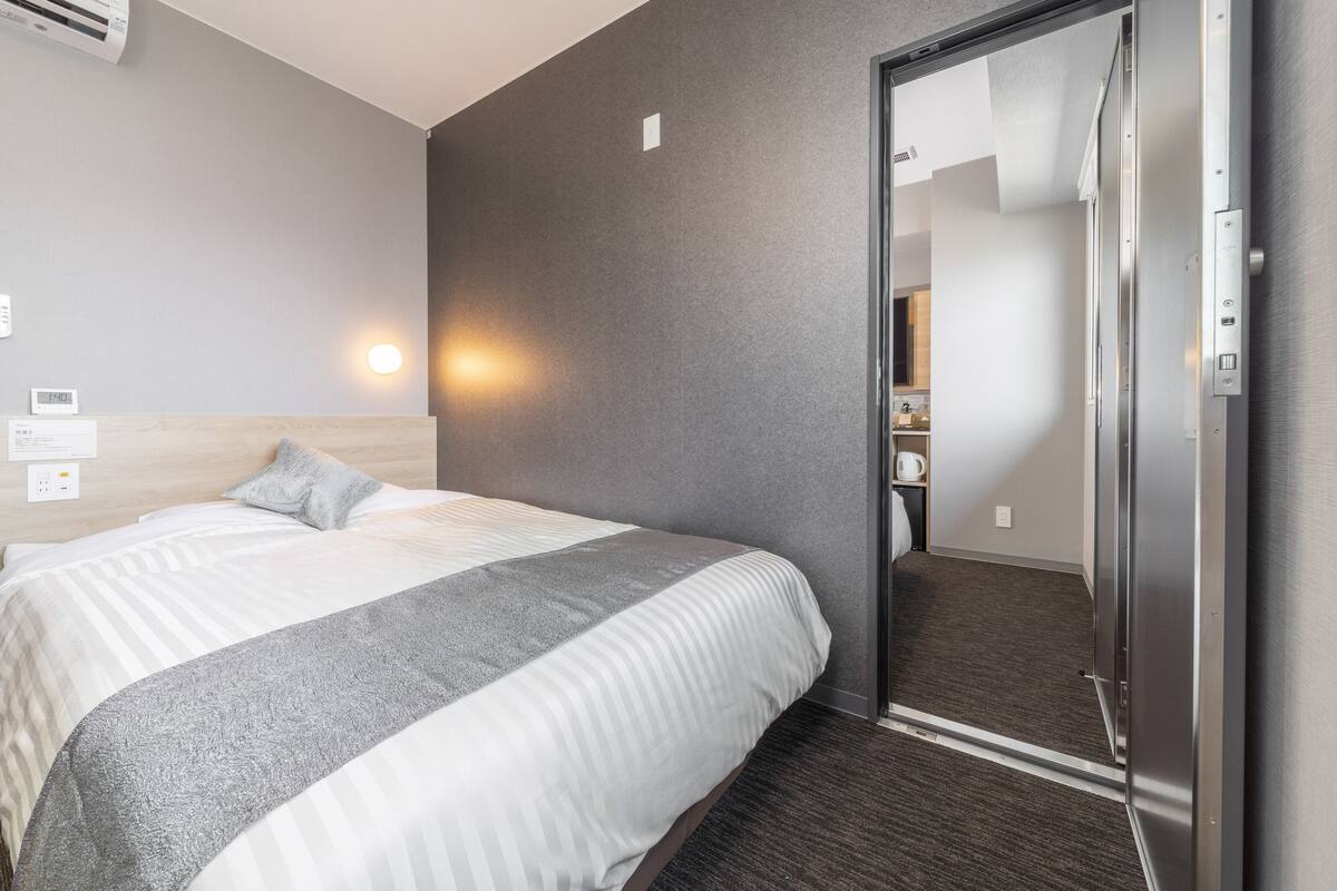 Una habitación de hotel con cama, aire acondicionado de pared, mesita de noche, espejo y puerta que da a un baño.