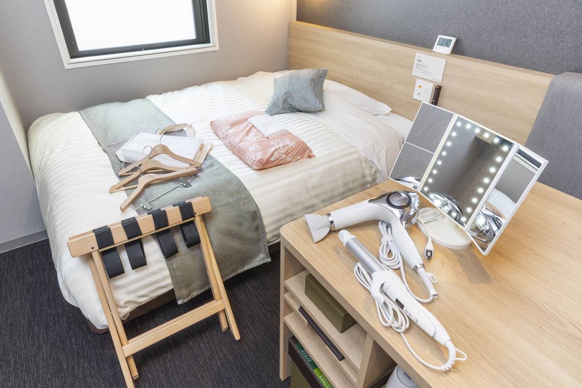 Habitación de hotel con cama, tocador con espejo e iluminación, escritorio con teléfono y secador de pelo, y ventana con vistas.
