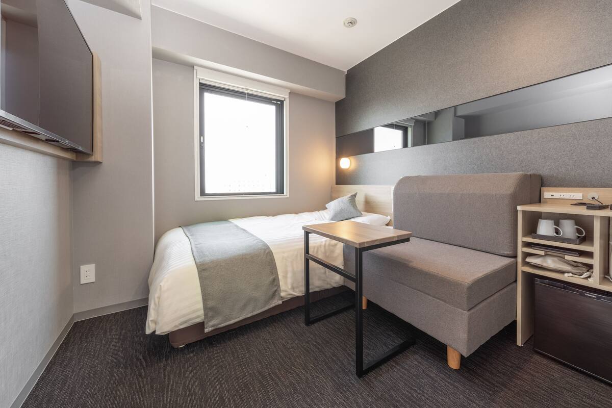 Habitación de hotel moderna con cama, escritorio pequeño, silla, televisor, ventana y estante empotrado con cafetera.