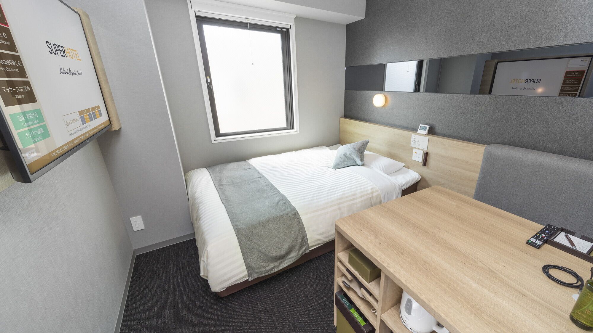 A compact hotel room with a bed, bedside table, and a small desk with a kettle.