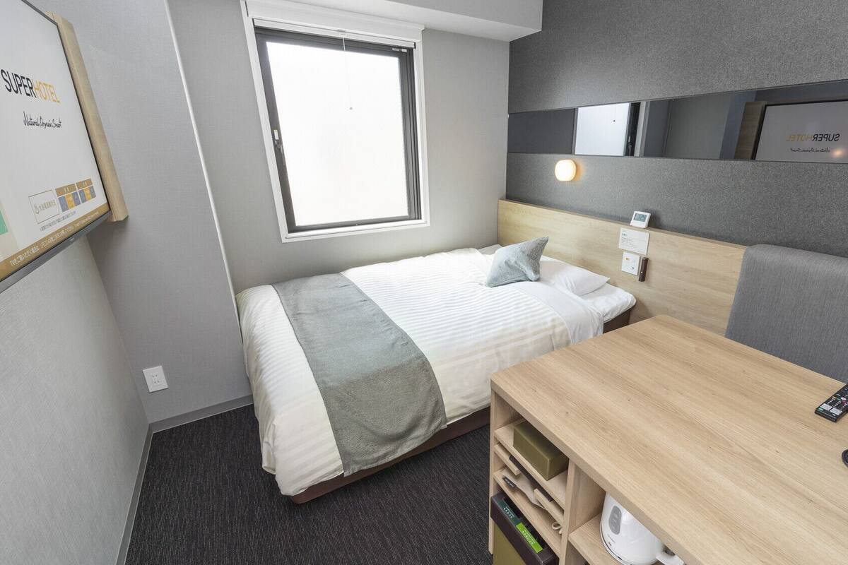 Habitación de hotel compacta con cama, mesita de noche y un pequeño escritorio con hervidor.