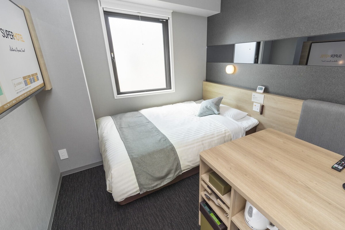 A compact hotel room with a bed, bedside table, and a small desk with a kettle.
