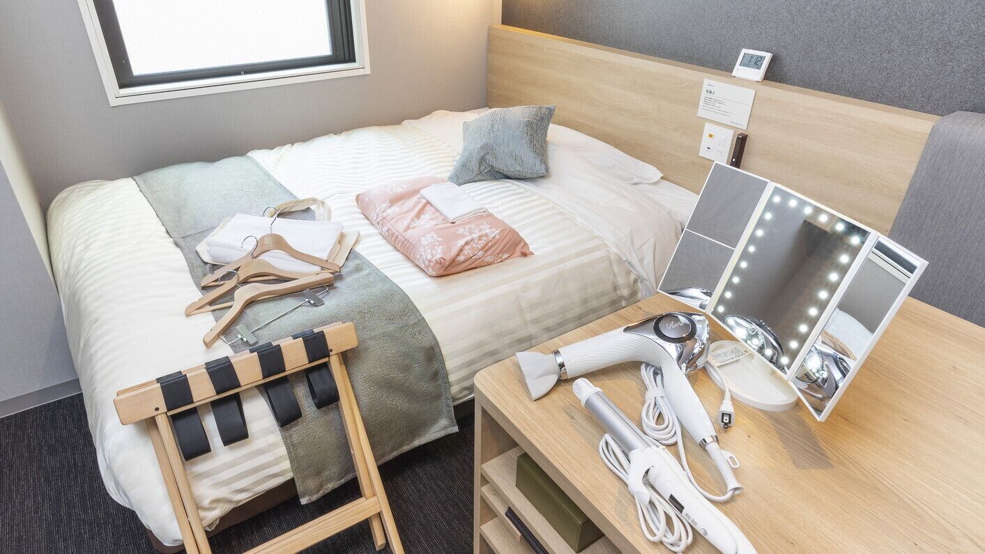 A neatly arranged hotel room with a bed, a bedside table with a mirror and lights, a chair, and a small desk with a hairdryer and other items.