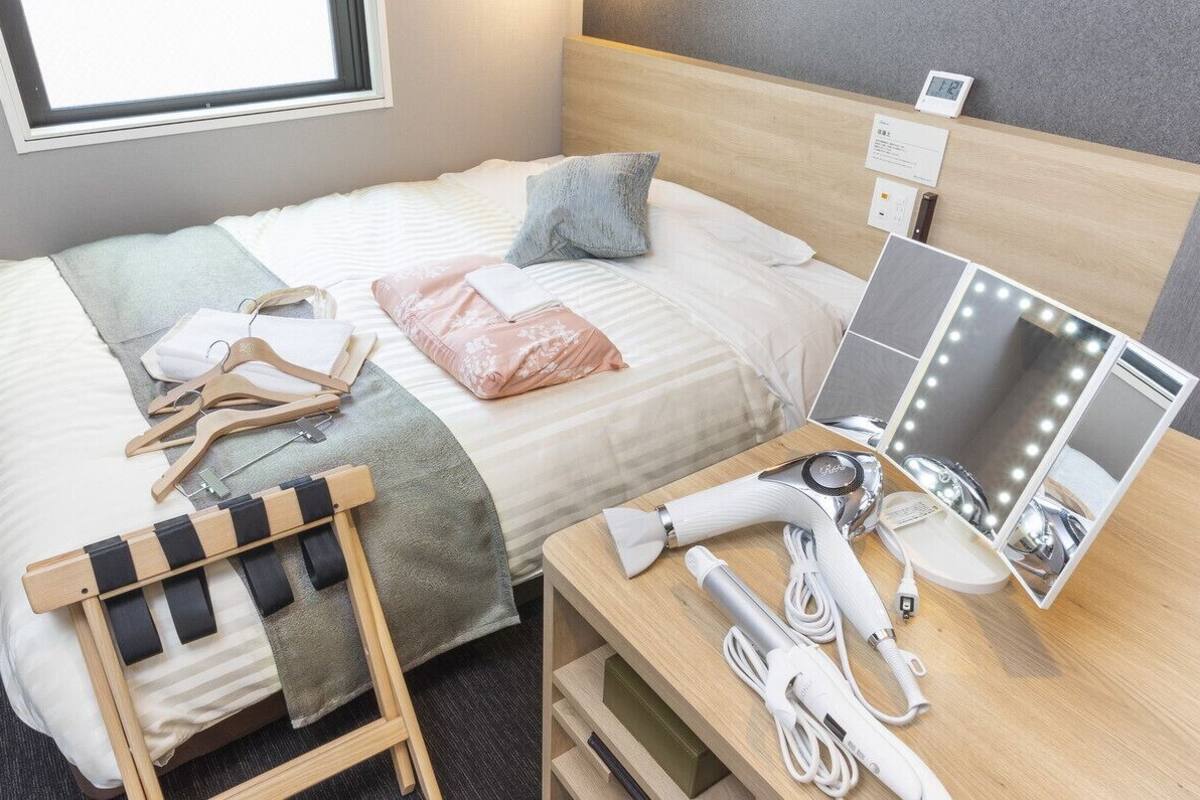 Habitación de hotel ordenada, con una cama, una mesita de noche con espejo e iluminación, una silla y un escritorio pequeño con un secador de pelo y otros objetos.