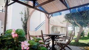 Eine Terrasse im Freien mit Tisch und Stühlen, umgeben von viel Grün und einem überdachten Bereich mit Blick auf ein Gebäude.