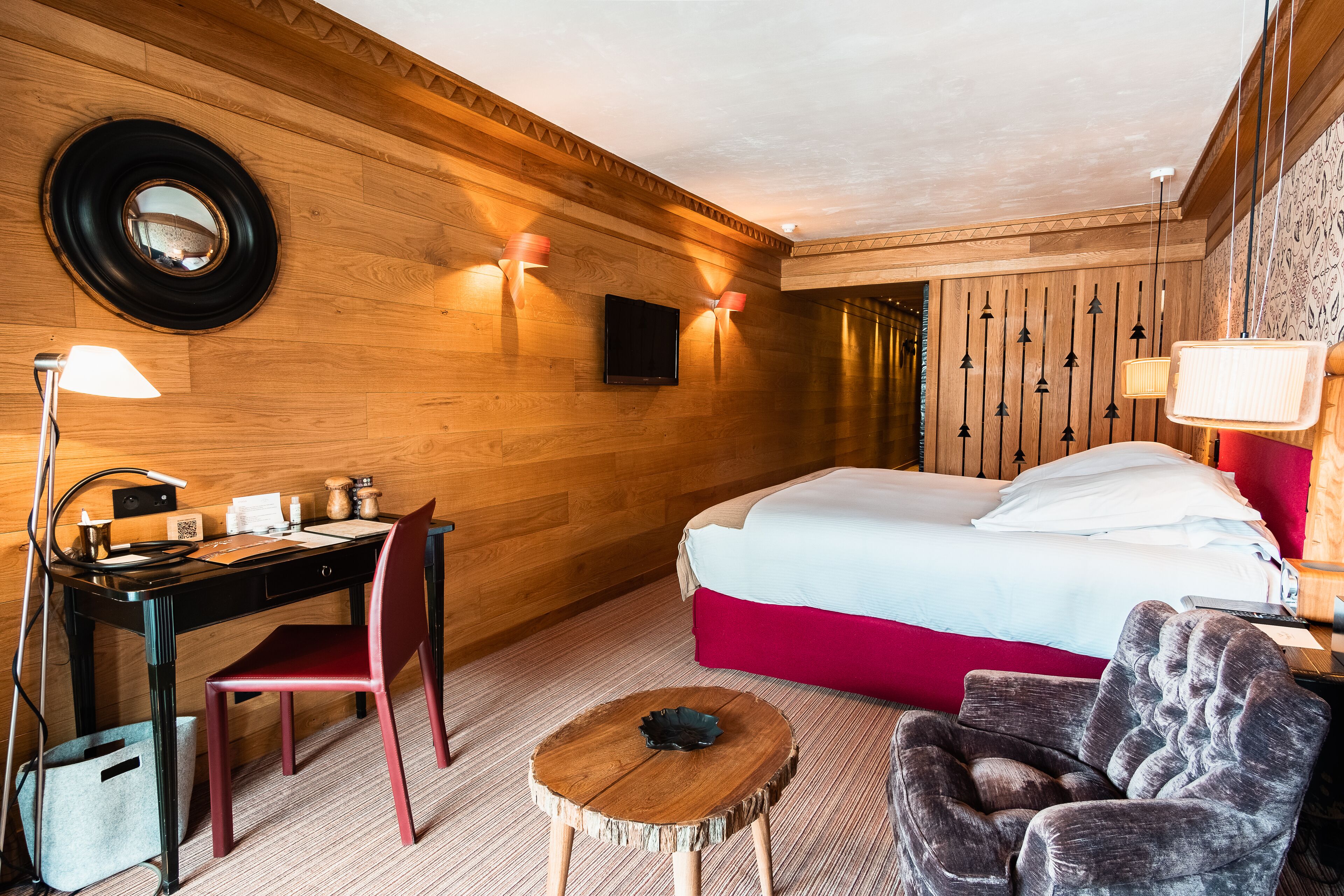 A hotel room with a wooden wall, a bed with a red accent, a desk with a chair, a small round table, and a TV mounted on the wall.