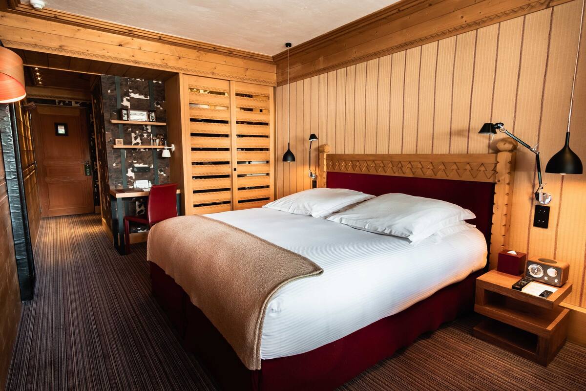 A hotel room with a large bed, wooden headboard, bedside lamps, a small table, and a red chair.